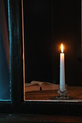 Wall Mural - Burning candle and Bible on wooden table at night, view through window