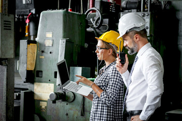 Young beautiful woman industrial engineer worker with helmet and safety glasses using laptop computer during discussing with senior businessman manager, people work at manufacturing industry factory