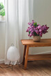 A bouquet of lilacs in a ceramic vase on an oak bench, a paper lamp on the floor by the window in the living room