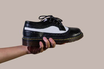 Wall Mural - A man's hand holds a black and white brogue wingtip shoe with a rubber outsole made from genuine cowhide. Men's hands holding elegant and shiny two tone shoes on a beige background