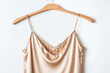 Smooth, sensual, beige color satin fabric woman's camisole on a clothes hanger, women's lingerie clothing product isolated on white background. Sexy, feminine, silky soft undergarment wear for ladies.