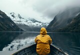 Fototapeta  - A contemplative moment captured of a lone person in a yellow raincoat, sitting in a boat facing a misty mountain landscape across serene waters