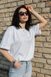 Cool beautiful fashionable woman model with cool sunglasses with curly hairstyle in a fashion white T-shirt stands near an antique brick wall