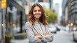 © Nataliia_Trushchenko - A woman is standing on a city street, smiling and wearing a gray jacket. She is crossing her arms and looking directly at the camera. Young happy pretty smiling professional business woman