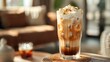 A tall glass of cold ice coffee with cream on top and brown liquid underneath, sitting on a table in front of an out of focus living room with soft lighting