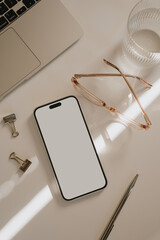 Office workspace table with mobile phone, laptop computer, glass of pure water, glasses. Soft sunlight glare and shadows. Aesthetic home office workspace. Blank white screen mock up with copy space