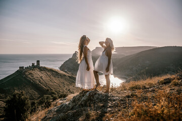 Wall Mural - Two young girls are standing on a hillside, one of them wearing a white dress. The sun is shining brightly, creating a warm and inviting atmosphere. The girls seem to be enjoying their time together.