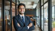 © Anzhela - Smiling confident young professional Indian businessman in dark suit standing with crossed arms the background of blurred office