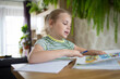 © evgeniia_1010 - A cute little girl does her homework with the help of a textbook, makes an entry in a workbook. The concept of online learning