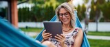 Fototapeta  - Happy woman reading on tablet while relaxing in a hammock outdoors.