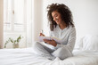 © Prostock-studio - A Hispanic woman is seated on a bed, focused on writing in a notebook. She appears engrossed in her writing, pen in hand, surrounded by a cozy bedroom setting.