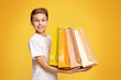 Little shopper. Happy teen boy holding lots of colorful paper shopping bags in hands, orange studio background