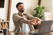 © Prostock-studio - Cheerful man is seated at a wooden desk in a room, taking a break from work to stretch his arms. He sits in front of an open laptop, suggesting he has been working and is now relaxing his muscles