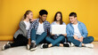 © Prostock-studio - Four multiethnic teenagers are casually seated on the floor with their backs to a vibrant yellow wall. One of them is holding an open laptop, showing something interesting