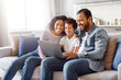 © Prostock-studio - African American family consisting of a father, mother, and two children sitting comfortably on a couch. They are all focused on a laptop screen, engaged in an activity together.