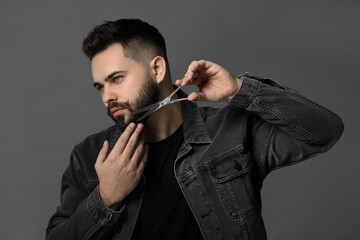 Wall Mural - Handsome young man trimming beard with scissors on grey background