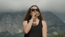 The Brunette's Long Hair Blows In The Wind As She Drinks Champagne Against The Backdrop Of Mountains In Clouds.