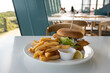 Close-up of a classic meal of a fried fish burger with some chips, a slice of lemon and dipping sauce on a table in a casual restaurant in the blurry background