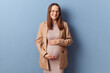 Beautiful delighted Caucasian young adult pregnant woman wearing dress and jacket posing isolated over blue background stroking her belly smiling to camera
