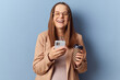 Cheerful joyful positive young adult pregnant woman wearing dress and jacket posing isolated over blue background using cell phone laughing reading funny news in internet
