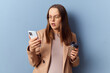 Shocked scared astonished Caucasian young adult pregnant woman wearing dress and jacket posing isolated over blue background reading surprised news in internet via smartphone