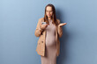 Confused puzzled young adult pregnant woman wearing dress and jacket holding mobile phone looking at smartphone display with uncertain posing isolated over blue background