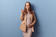 Delighted optimistic satisfied young adult pregnant woman wearing dress and jacket posing isolated over blue background holding smartphone looking at camera using internet