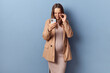 Serious concentrated young adult pregnant woman wearing dress and jacket posing isolated over blue background taking off her glasses looking at smartphone display reading message