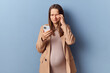 Sad tired young adult pregnant woman wearing dress and jacket posing isolated over blue background rubbing painful eyes after long hours using smartphone grimacing from unpleasant feelings