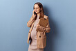 Beautiful young adult pregnant woman wearing dress and jacket posing isolated over blue background holding documents talking on smartphone with business partner