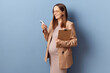 Pretty attractive young adult pregnant businesswoman wearing dress and jacket holding smartphone communicating online scrolling web pages posing isolated over blue background