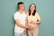 Loving young married pregnant family standing isolated over light green background man and woman hugging each other laughing stroking big belly enjoying pregnancy together