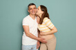 Smiling woman kissing husband cheek pregnant Caucasian young couple in casual clothing standing isolated over light green background loving family expecting baby