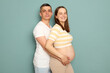 Happy cheerful satisfied pregnant Caucasian young couple standing isolated over light green background man embracing woman stroking belly couple looking at camera with smiles