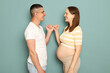 Cheerful young married pregnant family standing isolated over light green background holding fingers promises not fighting and quarrel looking at each other with love
