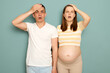 Shocked surprised displeased pregnant Caucasian young couple standing isolated over light green background touching heads making facepalm gestures saying omg