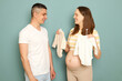 Young married pregnant family preparing for childbirth future mother showing to dad new tiny bodysuits buying for expecting kid standing isolated over light green background