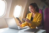 Fototapeta  - Young Asian businesswoman holding credit card and smartphone in online shopping using website on laptop sitting near window on airplane during flight, travel and business tourism concept.