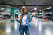 Young woman wearing blue jacket, standing with blurred car in parking lot. Beautiful female in underground parking with cars while looking for her car at night. lost car