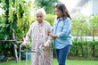 Caregiver help Asian elderly woman disability patient walk with walker in park, medical concept.