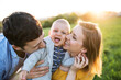 New parents holding small toddler, baby, outdoors in spring nature. Making baby laugh, kissing him on cheeks