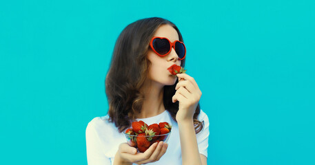 Wall Mural - Portrait of beautiful young woman with handful of fresh strawberries blowing kiss on blue background