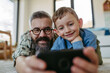 Little boy making selfie on smartphone with father, lying on floor in kids room, making silly faces. Dad explaining technology to son, digital literacy for kids.