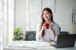A woman is sitting at a desk with a red coffee mug in front of her. She is smiling and she is enjoying her coffee. The scene suggests a relaxed and comfortable atmosphere