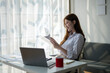 © Wasana - A woman is sitting at a desk with a laptop and a piece of paper. She is smiling and she is enjoying her work