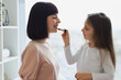© sofiko14 - The daughter paints lips to her beautiful mommy. Little girl doing makeup to mom sitting at bedroom. Happy Mother and daughter having fun together at weekend. Happy loving family.