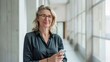 © Christian - smiling middle aged woman in business casual attire, looking at the camera, background features modern office building, copy and text space, 16:9