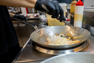 Poster - A chef in black gloves stir-fries noodles with vegetables in a stainless steel pan in a professional kitchen