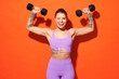 Young fitness trainer instructor woman sportsman wear purple top clothes spend time in home gym hold dumbbells raise up hands wink isolated on plain orange background. Workout sport fit abs concept.