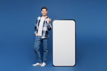 Full body young happy man he wear shirt white t-shirt casual clothes point finger on big huge blank screen mobile cell phone smartphone with area isolated on plain blue background. Lifestyle concept.